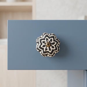 May include: A decorative cabinet knob with a floral design. The knob features a black and white pattern and a gold-colored center. It is mounted on a blue cabinet door.
