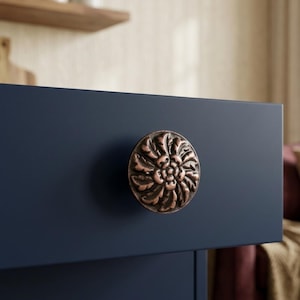 May include: A close-up of a dark blue cabinet drawer with a decorative, round, bronze-colored knob. The knob features a floral design with detailed leaf patterns. The cabinet is in a room with a neutral background.