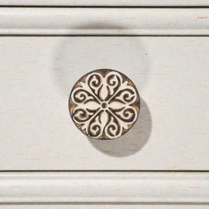 May include: A round, decorative cabinet knob with a floral design. The knob features a cream-colored pattern on a dark brown background. The knob is mounted on a white painted wooden surface.