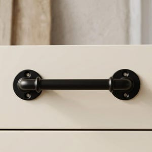 May include: A black metal cabinet pull with a cylindrical handle and circular mounting plates. The hardware is painted black and has a matte finish. The pull is mounted on a cream-colored cabinet door.