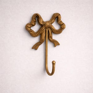May include: A gold-colored metal wall hook shaped like a bow. The bow is detailed with textured folds and a long, slender hook extending downwards. The hook has a rounded end. The wall hook is mounted on a white surface.