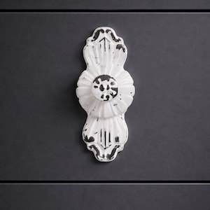 May include: White distressed decorative cabinet pull with a floral design. The pull is mounted on a dark gray cabinet door. The pull has a central circular element and elongated, ornate ends.
