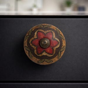 May include: A round, decorative cabinet knob with a floral design. The knob features a red flower at the center, surrounded by brown and green leaf accents. The knob is mounted on a black surface.