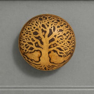 May include: A round, wooden decorative element featuring a detailed tree of life design. The tree is etched in dark brown against a light brown background. The item is circular and appears to be a decorative accent.