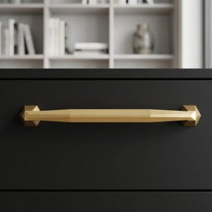 May include: A gold-colored, geometric drawer pull is mounted on a black drawer. The pull has a faceted design with hexagonal end pieces and a rectangular center. The background shows a white bookshelf with books and decorative items.