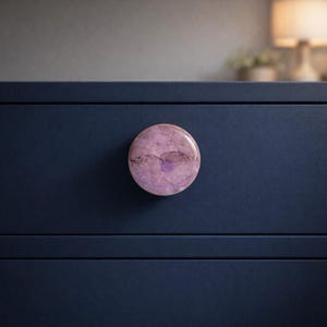 May include: A round cabinet knob with a marbled appearance in shades of pink and purple. The knob is mounted on a dark blue cabinet door. The knob's design features a glossy finish, adding a touch of elegance to the furniture.