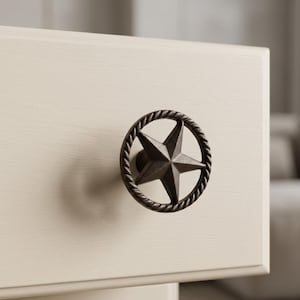 May include: A bronze-colored star-shaped drawer pull with a rope-like border is mounted on a cream-colored cabinet. The star has five points and is centered within the circular border. The hardware adds a rustic touch.