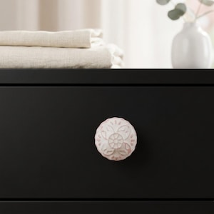 May include: A close-up of a black drawer with a white and pink floral ceramic knob. The knob is round with a detailed embossed design. Beige folded linens are stacked on the drawer above.