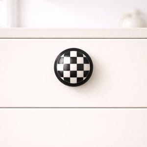 May include: A round cabinet knob with a black and white checkered pattern. The knob has a black border and is mounted on a white drawer. This decorative knob adds a unique touch to furniture.