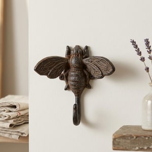 May include: A cast iron bee-shaped wall hook with a dark brown finish. The hook features detailed wings and body, and a single hook extending downwards. The decorative item is mounted on a white wall.