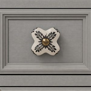 May include: A decorative cabinet knob with a white base and black floral design. The knob has a brass-colored center and is mounted on a gray cabinet door. The knob is shaped like a four-leaf clover.