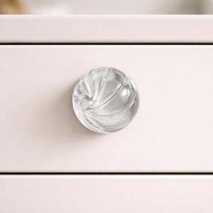 May include: A clear, glass cabinet knob with a spiral design. The knob is round and has a textured surface. It is mounted on a light pink cabinet drawer.