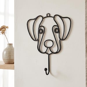 May include: Black metal dog-shaped wall hook. The hook is designed in a minimalist style, featuring the outline of a dog's face with large ears and a single hook at the bottom. It is mounted on a white wall, suitable for coats, keys, or other items.