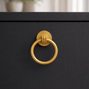May include: Close-up of a gold-colored ring pull hardware on a black cabinet. The ring pull is circular with a textured finish and attached to a round backplate. The hardware adds a touch of elegance to the furniture.