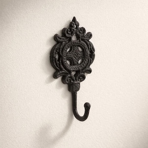 May include: Ornate black cast iron wall hook. The hook features a decorative, scrolled design with a central circular element and a curved hook. The hook is mounted on a white wall, showcasing its intricate details and vintage aesthetic.
