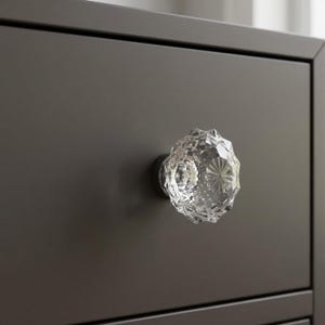 May include: Close-up of a clear, faceted crystal knob on a dark brown drawer. The knob has a round, starburst design in the center and is mounted on a dark metal base. The drawer is part of a larger piece of furniture.