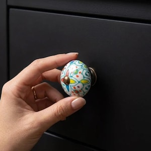May include: A decorative ceramic cabinet knob with a white base and colorful floral pattern. The knob features a brass-colored metal base and a small brass-colored center accent. The knob is held against a black cabinet.