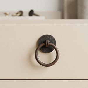 May include: Close-up of a bronze-colored ring pull hardware attached to a cream-colored cabinet drawer. The ring pull has a circular backplate and a round handle. Other hardware is visible in the background.