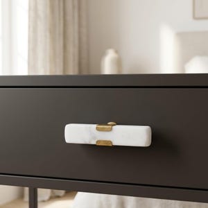 May include: Close-up of a black drawer with a white marble and brass handle. The rectangular handle has a polished finish and is mounted on the drawer front. The drawer is part of a larger piece of furniture.