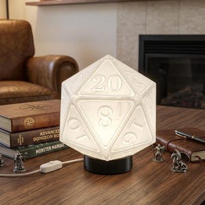 May include: A glowing white D20 lamp with a black base sits on a wooden table. The lamp is lit, displaying the numbers 1 through 20. Miniature figurines and books with titles like "Advanced Dungeon Ruin" and "Monster Manual" are also on the table.