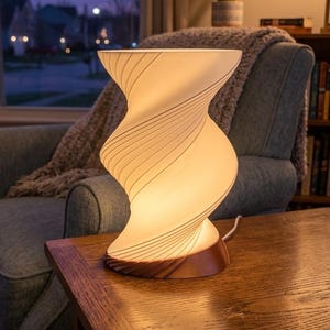 May include: A decorative table lamp with a unique, swirling design. The lamp has a white, frosted glass shade with thin, grey lines and a wooden base. The lamp is lit, casting a warm glow on the wooden table.