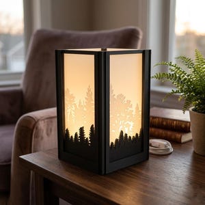 May include: A black, rectangular lamp with a forest silhouette design. The lamp has a warm, glowing interior and a white power cord. The lamp is sitting on a wooden table. The lamp's design features trees and a sunset.