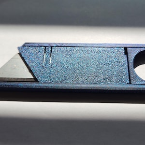 Könnte beinhalten: Ein blaues Teppichmesser mit strukturierter Oberfläche und silberner Klinge. Das Messer hat einen abgerundeten Griff mit einer kreisförmigen Öffnung und einer verschiebbaren Klingenabdeckung. Die Klinge ist teilweise ausgefahren.