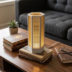 Puede incluir: Una lámpara de mesa hexagonal moderna con un diseño de rejilla, que emite un brillo cálido. La lámpara blanca está sobre libros en una mesa de centro de madera. Una pequeña planta suculenta en maceta y un diario encuadernado en cuero también están sobre la mesa.