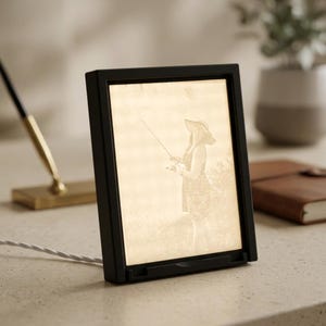 May include: Black rectangular photo frame with a sepia-toned image of a person fishing. The frame has a built-in stand and a white cord. A pen and a notebook are visible in the background.