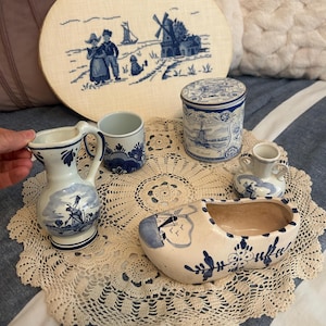 May include: A collection of blue and white ceramic items including a pitcher, a mug, a lidded canister labelled Holland, a small two-handled vase, and a clog-shaped planter, all featuring windmill motifs, alongside an oval embroidered wall hanging.