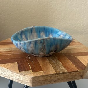 May include: A ceramic bowl with a unique, organic shape. The bowl features a marbled design with shades of blue, white, and gray. The bowl is displayed on a wooden table with a geometric pattern and black metal legs.
