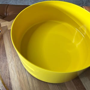 May include: A bright yellow, round bowl with a smooth, glossy finish. The bowl is empty and appears to be made of plastic or a similar material. It sits on a wooden surface with visible grain patterns. The bowl's interior reflects the light.