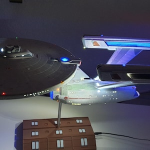 Könnte beinhalten: Ein detailliertes Modell des Raumschiffs Enterprise NCC-1701 mit einer großen kreisförmigen Untertassensektion, zwei länglichen Warpgondeln mit leuchtend blauem Licht und einem Sekundärrumpf, montiert auf einem rechteckigen braunen Sockel.