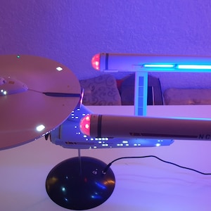 Könnte beinhalten: Ein Modell des Raumschiffs USS Enterprise mit einer weißen Untertassensektion, der Aufschrift NCC-1701, zwei langen zylindrischen Gondeln mit leuchtend blauem Licht und rot beleuchteten Spitzen, montiert auf einem schwarzen runden Sockel.