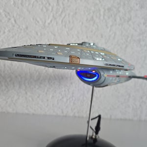 Może przedstawiać: Szczegółowy model statku kosmicznego USS Voyager z serii Star Trek, posiadający szary kadłub z beżowymi akcentami, podświetlane na niebiesko gondole warp oraz świecące na czerwono światła nawigacyjne, umieszczony na cienkim metalowym stojaku.