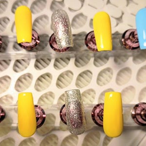 May include: A set of artificial nails in various colors, including light blue, yellow, and silver glitter. The nails are displayed on a white surface with a grid pattern, each nail is attached to a pink and silver holder.