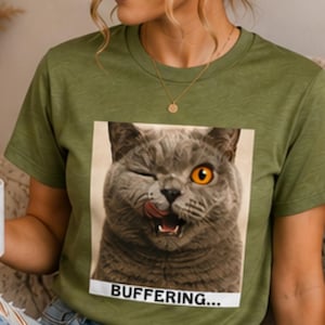 Può includere: Una t-shirt a maniche corte color verde oliva con una stampa grafica quadrata di un gatto grigio che strizza l'occhio e si lecca il naso, con un occhio arancione brillante visibile. Sotto l'immagine del gatto, il testo "BUFFERING..." è stampato in nero.
