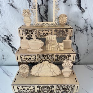 May include: A three-tiered wooden display stand featuring laser-cut designs related to Dia de los Muertos. The stand includes decorative panels with skulls, flowers, and the text Dia de los Muertos, along with separate wooden cutouts of skulls, a frame, a cup, candles, and bread.
