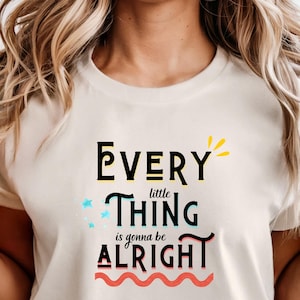 Op de afbeelding: Een ivoorkleurig T-shirt met korte mouwen en de tekst "Every little THING is gonna be ALRIGHT" in zwarte, blauwe en rode letters, geaccentueerd met kleine blauwe sterretjes, een gele zonnestraal en een golvende rode lijn.