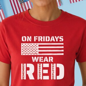 Może przedstawiać: Czerwona koszulka z napisem "ON FRIDAYS WEAR RED" i wzorem amerykańskiej flagi. Koszulka zawiera również słowa "REMEMBER EVERYONE DEPLOYED". Koszulka jest noszona przez osobę.