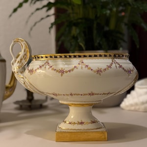 Könnte beinhalten: Eine weiße Porzellan-Servierschüssel mit goldenem Rand und Sockel. Die Schüssel hat eine dekorative Blumengirlande in Lila und Gold. Der Griff ist wie ein Schwanenkopf geformt. Die Schüssel ist etwa 25 cm breit und 15 cm hoch.