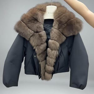 May include: A dark navy blue jacket with a large, plush brown fur collar and trim. The jacket has a cropped design with long sleeves and a zipper closure. The fur appears to be fox or similar, adding a luxurious touch to the outerwear.