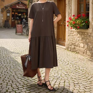 May include: A dark brown, short-sleeved dress with a tiered skirt, paired with black sandals. A woven tote bag with a patterned scarf is held. The dress has a simple neckline and is accessorized with a silver necklace. The setting is a cobblestone street.
