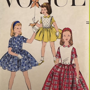 May include: Vintage Vogue pattern cover featuring illustrations of three young girls in dresses. The dresses are in blue floral, yellow, and red plaid patterns. The Vogue logo is at the top in black letters.