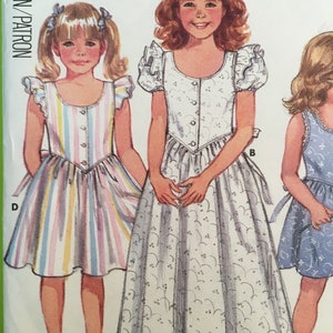 May include: A vintage sewing pattern featuring illustrations of three girls in dresses. One dress is striped in pastel colors, another is white with a floral pattern, and the third is blue with a floral pattern. The dresses have button closures and bows.