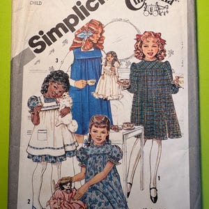 Pode incluir: Padrão de costura Simplicity Cinderella para um vestido infantil, tamanho 6. O padrão apresenta ilustrações de meninas em vários vestidos, incluindo um macacão azul, um vestido xadrez e um vestido floral. O padrão inclui instruções para diferentes vistas.
