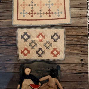 May include: Two rag dolls, one wearing a red and white dress and holding a watermelon, the other wearing a brown and white dress, sitting on a blue bench in front of two framed quilts with red, blue, and white patterns.