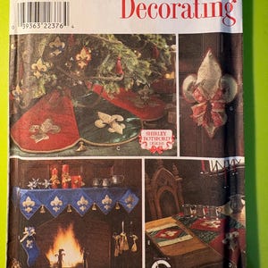 Op de afbeelding: Simplicity Home Decorating patroon met kerstontwerpen. De afbeelding toont een kerstboomrok, manteldecoraties en een tafelloper, allemaal versierd met fleur-de-lis patronen. Het patroon bevat de tekst "ONE SIZE".