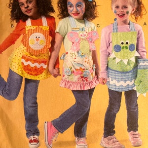 May include: Three children wearing colorful aprons with animal designs. The aprons have pockets and are decorated with appliques. The children are wearing jeans and sneakers. The aprons are labeled A, B, and C.