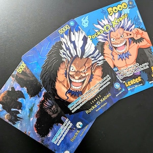 May include: Three One Piece trading cards featuring the character Rocks D. Xebec. The cards display a blue background, the text Rocks D. Xebec, a power level of 5000, and the label Leader. The artwork depicts a character with spiky hair.